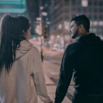 A couple holding hands and walking down a city street at night, sharing a quiet, intimate moment together.